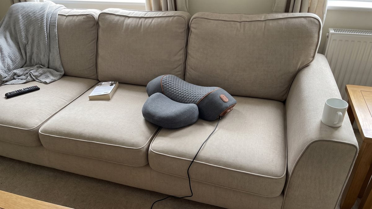 The FisioRest sitting askew on the middle cushion of a family sofa, its cable trailing off the front, a throw and a remote beside it.