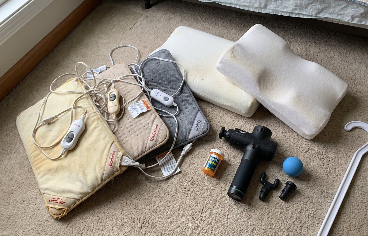 Six years of bought remedies laid out on a bedroom carpet: three heating pads of different ages, two contour pillows, a massage gun with spare heads, a lacrosse ball, a pill bottle and a long massage hook cut off by the edge of the frame.